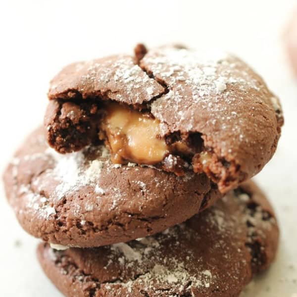 Two chocolate cookies stacked, one with a bite showing a gooey caramel center, lightly dusted with powdered sugar on a white surface.