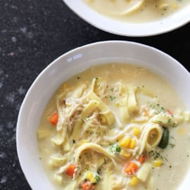 Two bowls of creamy chicken noodle soup with vegetables, including carrots, corn, and zucchini, are served on a dark surface.