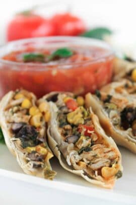 Close-up of sliced stuffed rolls filled with corn, beans, and vegetables on a plate. A bowl of chunky tomato salsa in the background.