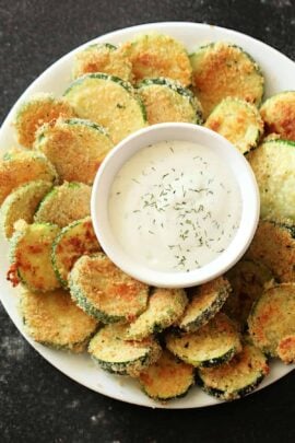 A plate of breaded and fried zucchini slices surrounding a small bowl of creamy dipping sauce garnished with herbs.