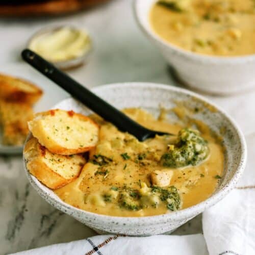 A ceramic bowl filled with creamy soup, featuring pieces of broccoli and bread slices, with a black spoon resting inside. Another similar bowl and a small dish of butter in the background.