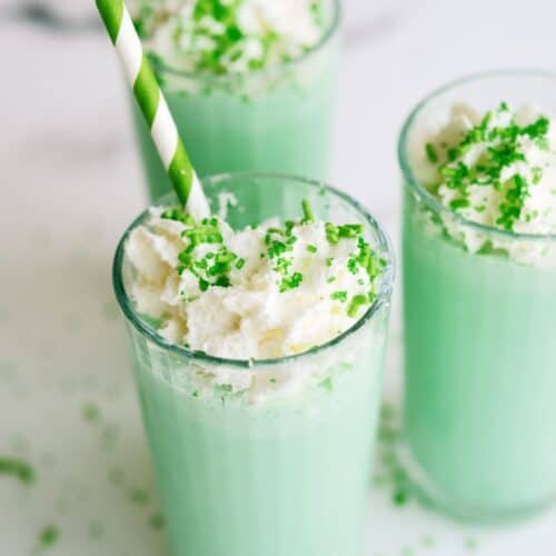 Copycat McDonald's Shamrock Shake in 3 glasses topped with whip cream and green sprinkles. Green and white striped straws in each glass.