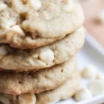 4 White Chocolate Macadamia Nut Cookies stacked on a baking sheet with white chocolate chips sprinkled around the bottom.