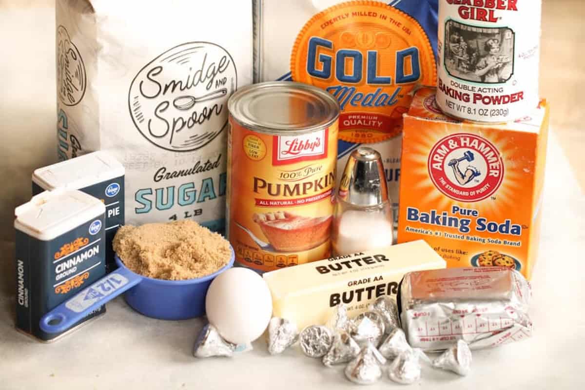 A collection of baking ingredients including flour, sugar, pumpkin, baking soda, baking powder, cinnamon, brown sugar, butter, salt, chocolate, and baking chocolate squares.