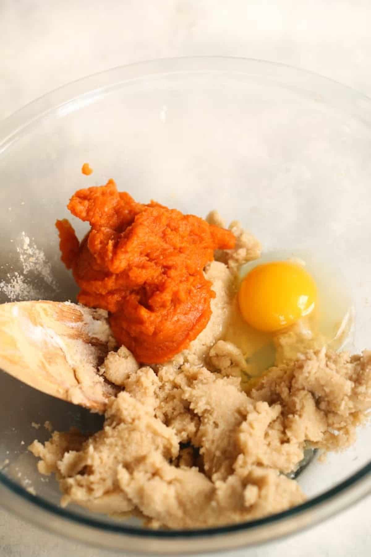 A clear mixing bowl contains pumpkin puree, an egg, and partially mixed brown sugar and butter. A wooden spoon is also in the bowl.