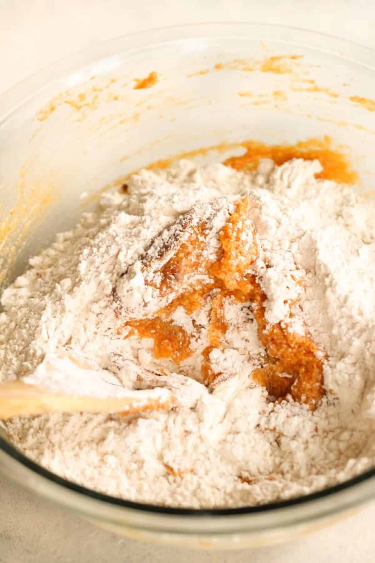 A glass bowl containing a mixture of flour and batter, being stirred with a wooden spoon.