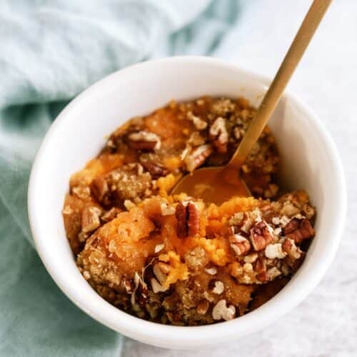 A white bowl filled with sweet potato casserole topped with pecans and a spoon in it on a cloth surface.