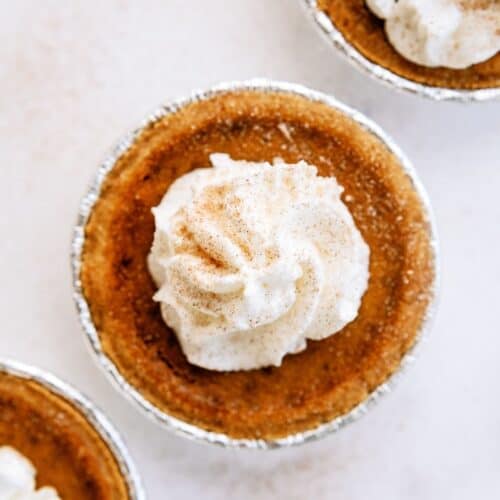 A small pumpkin pie topped with a swirl of whipped cream on a light background.