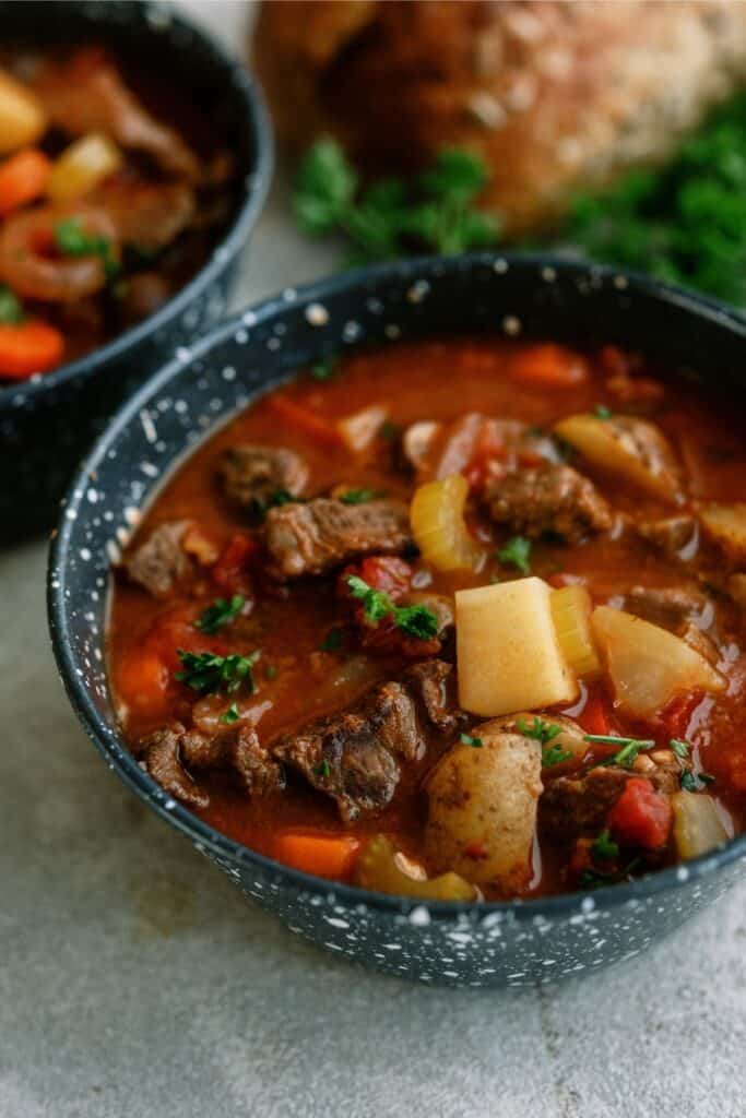 A bowl filled with hearty beef stew containing chunks of beef, potatoes, celery, carrots, and garnished with fresh parsley. Another bowl of stew and a loaf of bread are blurred in the background.