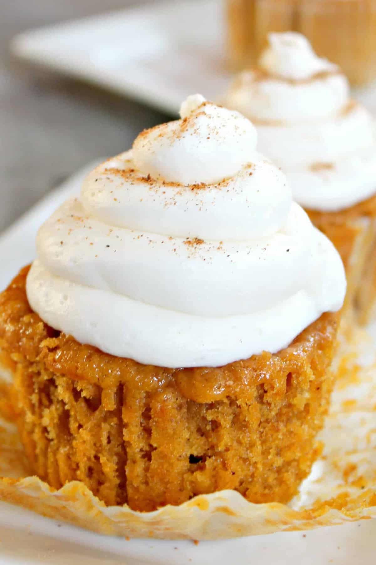 A close-up of a pumpkin cupcake with white frosting sprinkled with cinnamon on top. The partially unwrapped cupcake sits on a white plate.