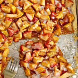 A piece of fruit tart with a combination of sliced apples and strawberries on a flaky crust and drizzled with icing on a baking sheet. A fork is placed beside the tart.