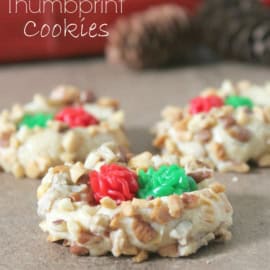 Three thumbprint cookies topped with green and red icing are displayed on a brown surface with pine cones in the background.