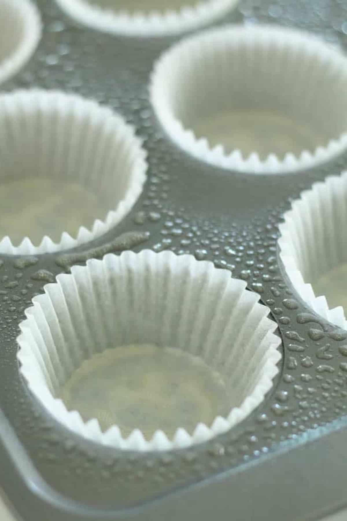 Close-up of a muffin tin with six paper baking cups inside, and droplets of water on the tin surface.