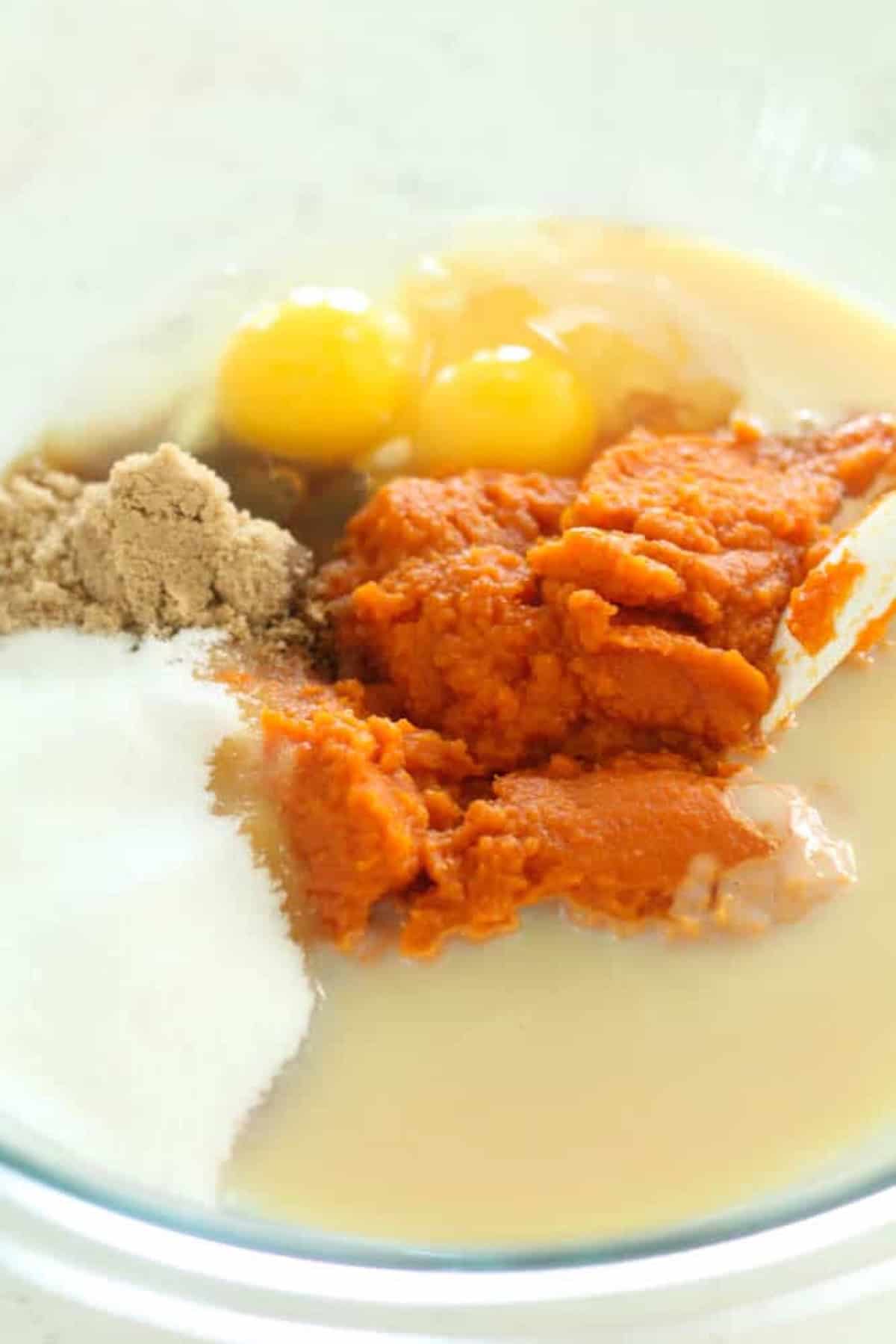 A mixing bowl with ingredients for pumpkin pie, including brown sugar, white sugar, pumpkin puree, eggs, and condensed milk.