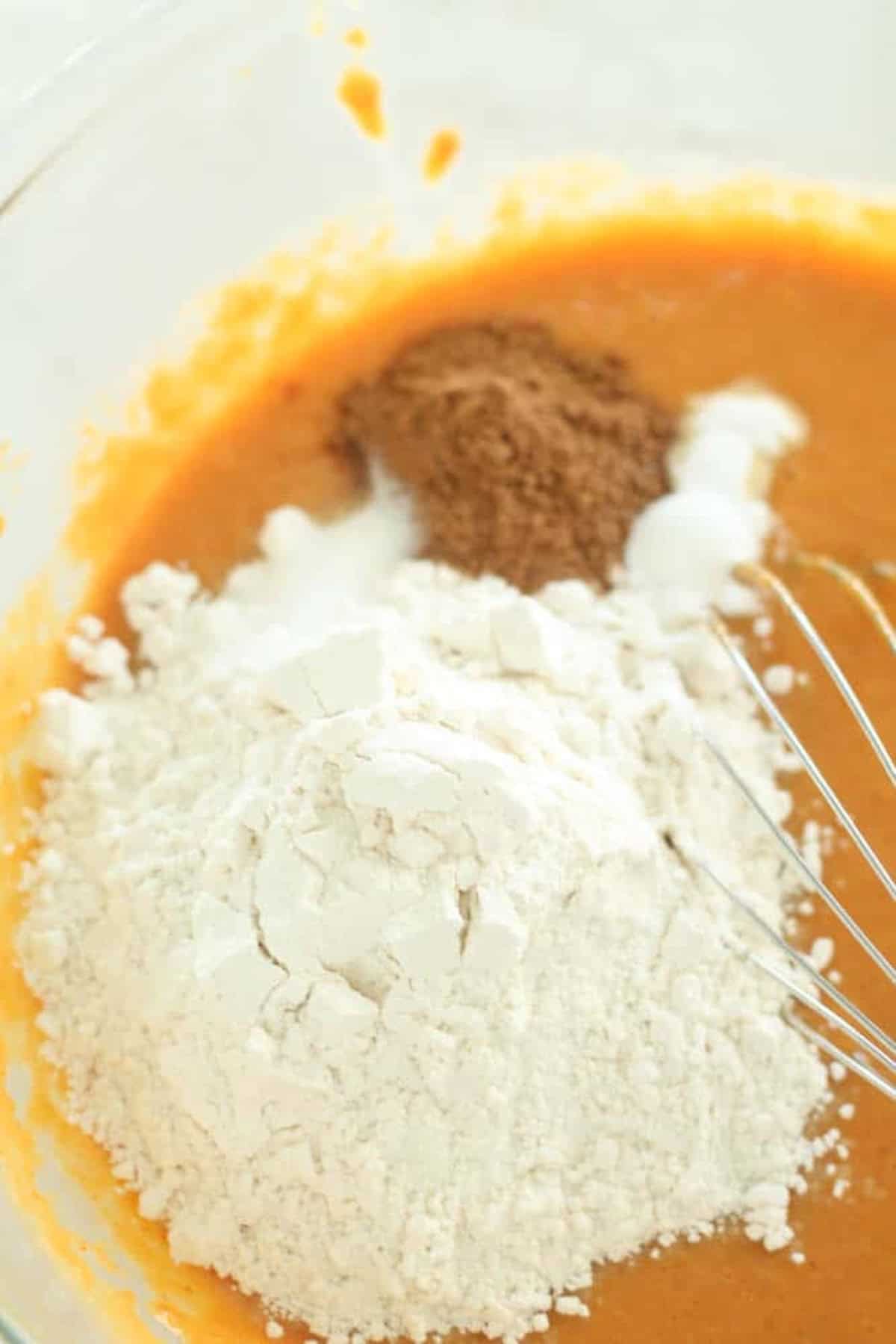 A clear bowl containing pumpkin puree, flour, sugar, and dark brown spices, with a wire whisk partially visible.