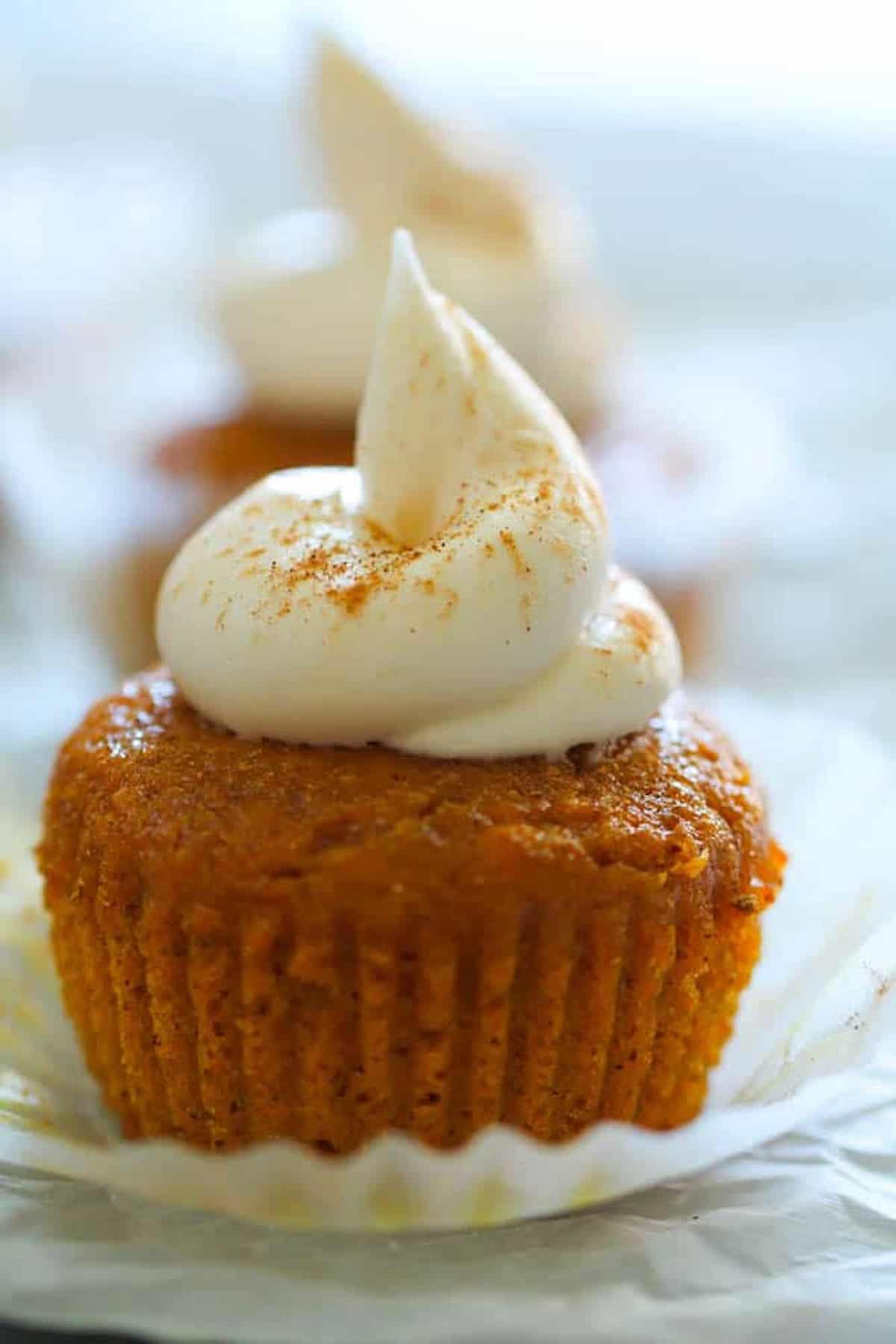 A close-up of a pumpkin cupcake topped with cream cheese frosting and a sprinkle of cinnamon, with another similar cupcake blurred in the background.