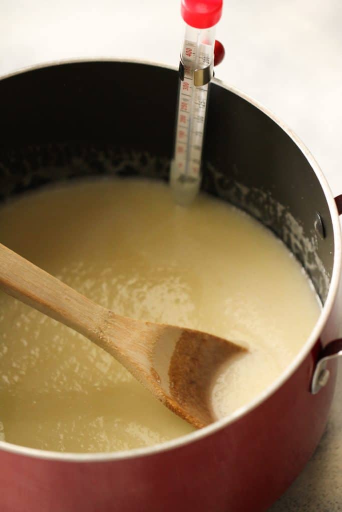 A large pot on the stovetop with a sugar mixture in it with a wooden spoon and a candy thermometer attached to the side.