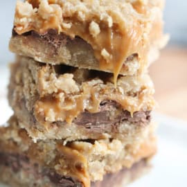 A stack of three caramel oat bars with gooey caramel and chocolate layers sits on a white plate.