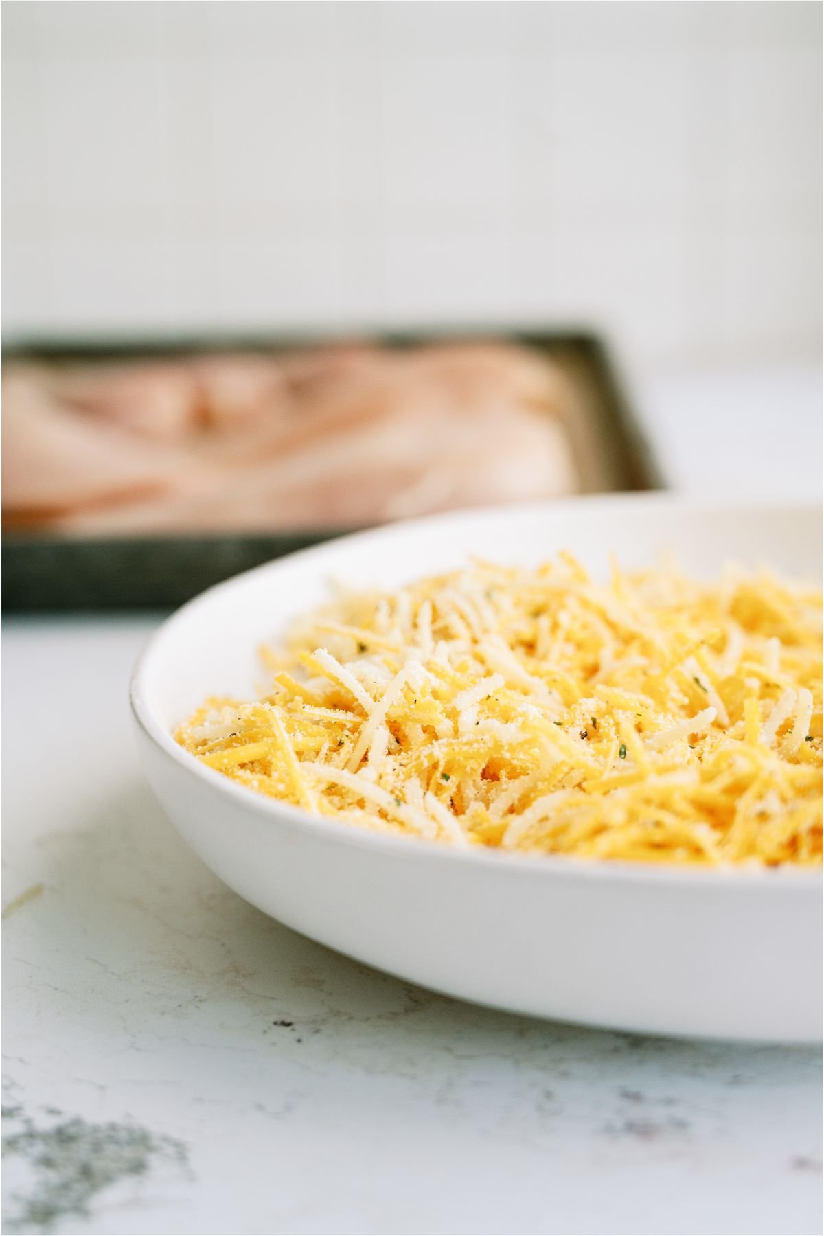 A bowl of shredded cheese and a pan with uncooked chicken breast in the background.