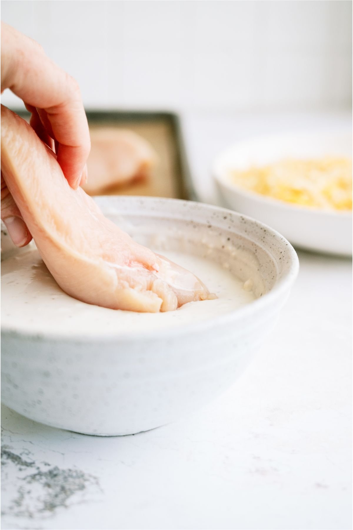 Dipping a raw chicken breast in the ranch mixture in the bowl. A bowl of cheese and a pan of uncooked chicken breasts in the background. 