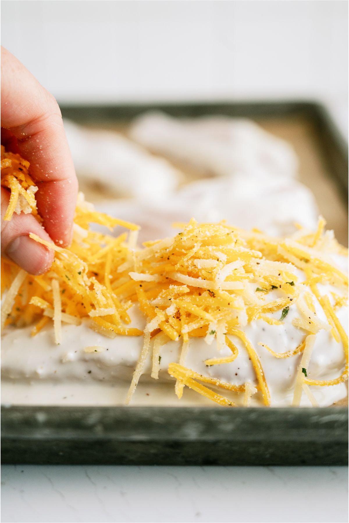 Sprinkling shredded cheese on top of dipped chicken breast on sheet pan.