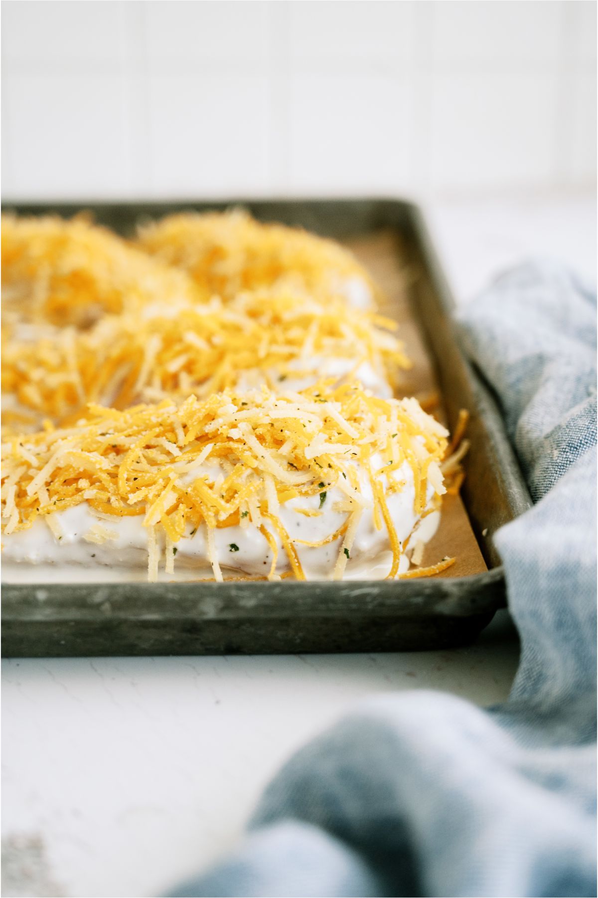 Several unbaked chicken breast dipped in ranch mixture and topped with cheese on a lined baking sheet. 