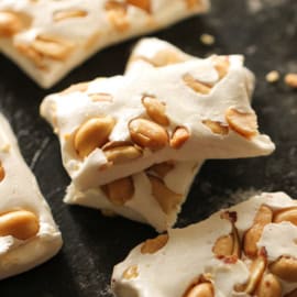 Close-up of nougat candy pieces containing peanuts, displayed on a dark surface.