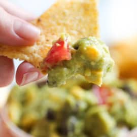 A hand holds a tortilla chip with a scoop of guacamole, containing chunks of tomato and corn. The background shows more guacamole.