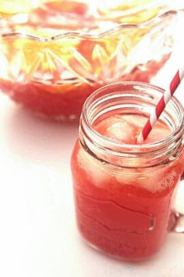 A mason jar filled with a red iced drink and a red-and-white striped straw. A glass pitcher filled with the same drink is in the background.