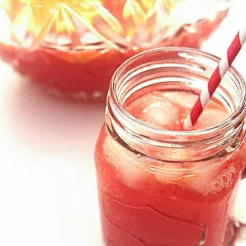 A mason jar filled with a red iced drink and a red-and-white striped straw. A glass pitcher filled with the same drink is in the background.