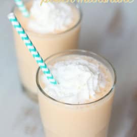 Two glasses of peach milkshake topped with whipped cream and served with green and white striped straws.