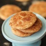 3 Snickerdoodle Cookies on top of a small cake stand with remaining Snickerdoodle Cookies in the background.