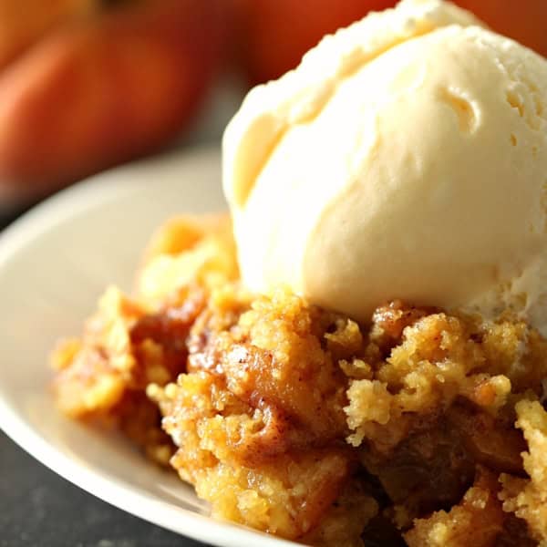 A serving of peach cobbler topped with a scoop of vanilla ice cream on a white plate, with two whole peaches in the background.