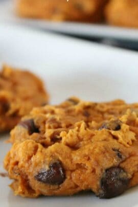 A close-up of a chocolate chip cookie with a soft, doughy texture on a white surface. Another cookie is blurred in the background.