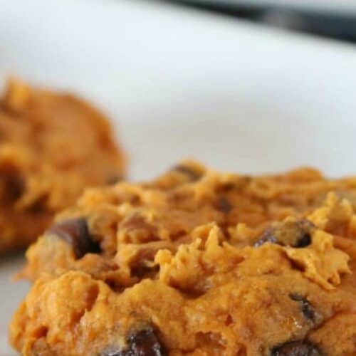 A close-up of a chocolate chip cookie with a soft, doughy texture on a white surface. Another cookie is blurred in the background.