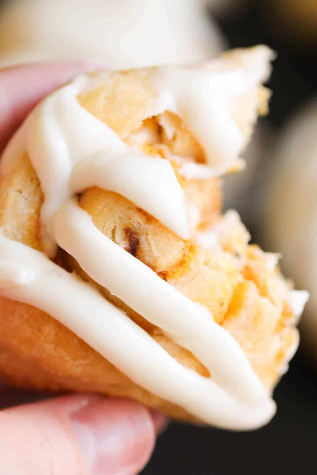 Close-up of a person's hand holding a small piece of pastry with white icing drizzled on top. The pastry appears to be partially eaten.