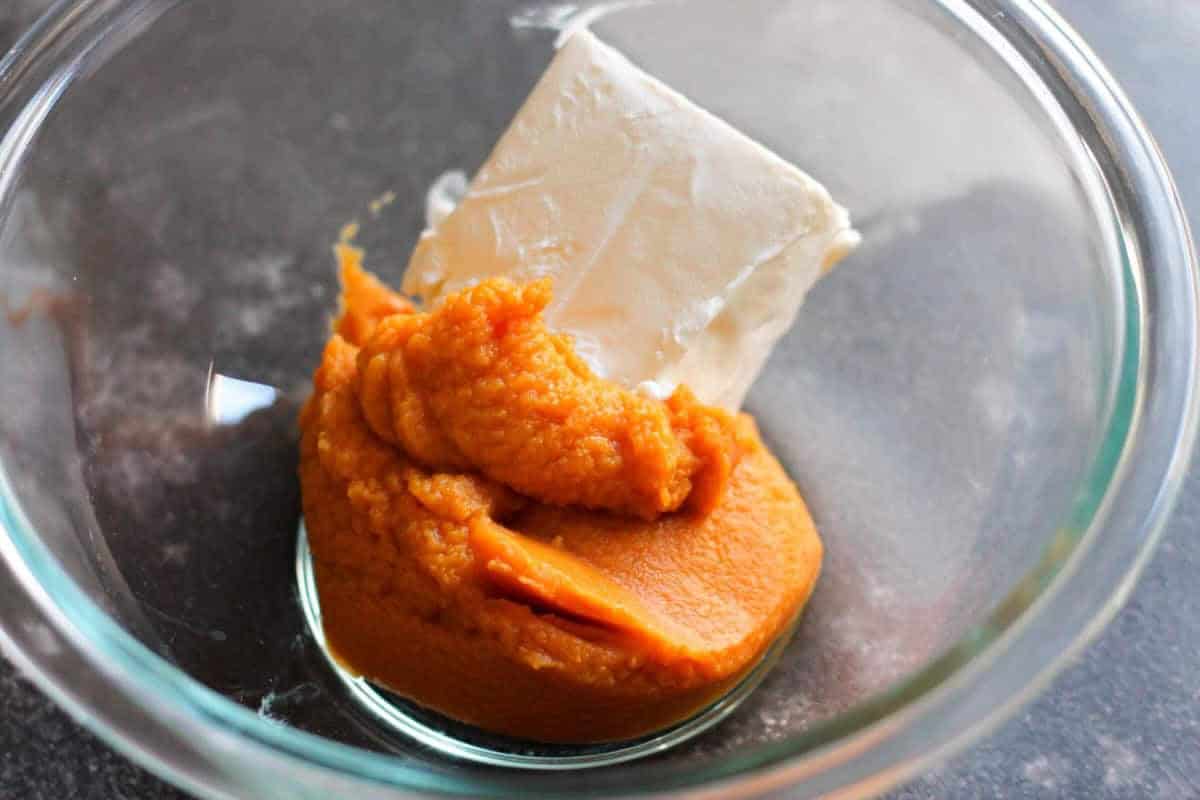 A glass bowl containing a block of cream cheese and a portion of pumpkin puree.