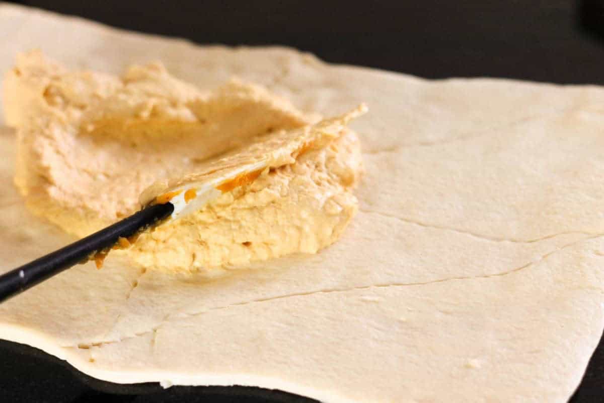 A utensil spreads a creamy mixture onto an uncooked rectangular piece of dough.