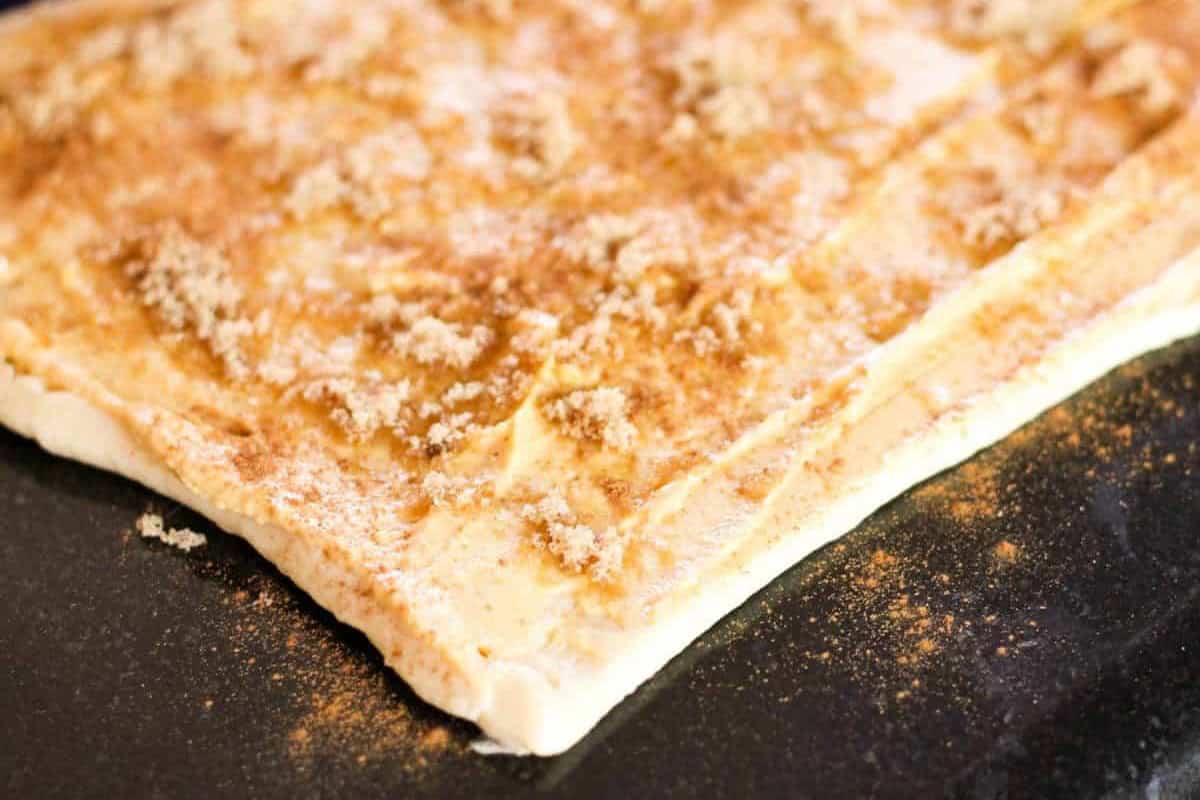 A sheet of dough is covered with a sprinkle of brown sugar and cinnamon on a dark surface, ready for baking.