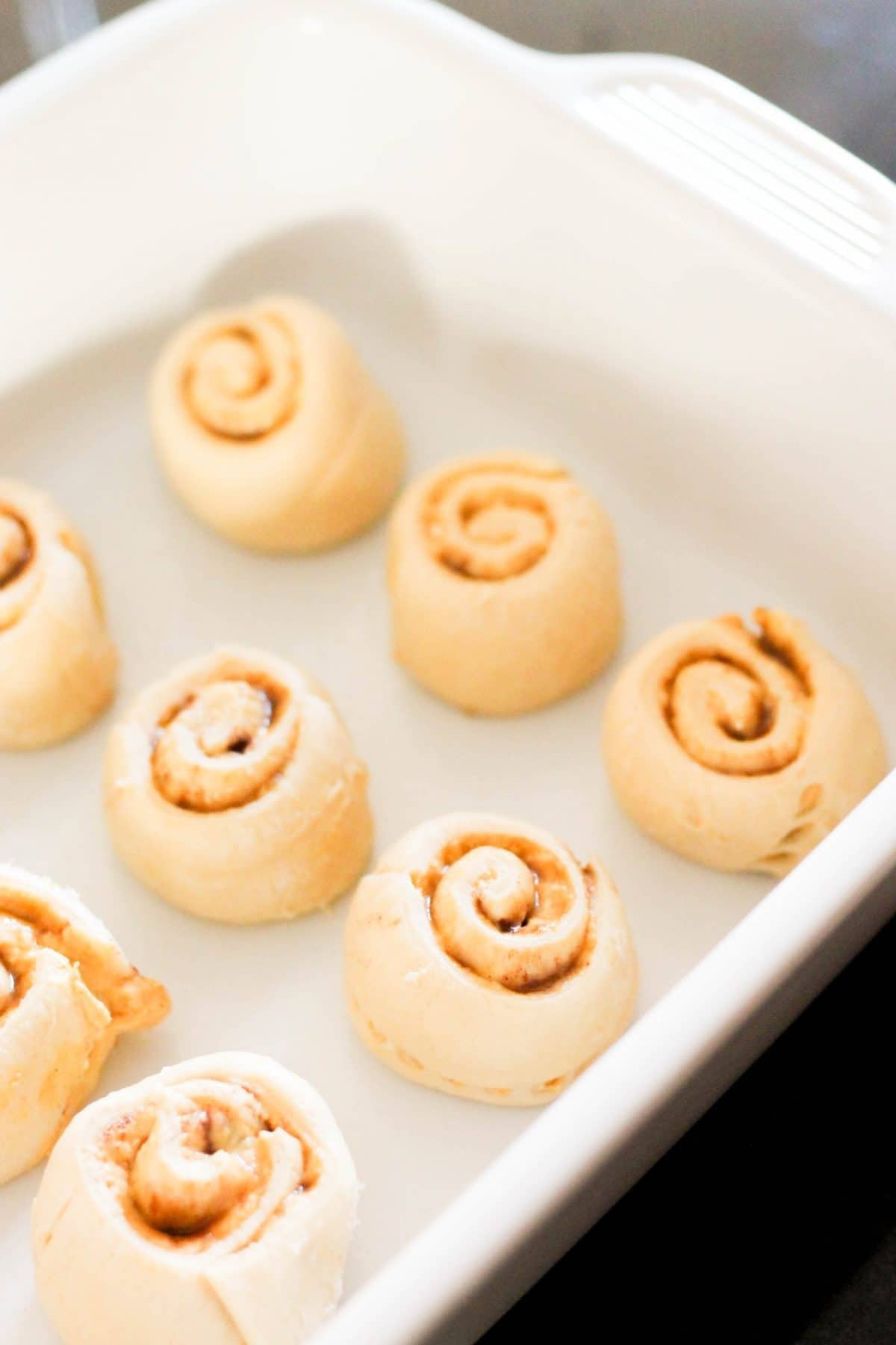 A white baking dish contains seven uncooked cinnamon rolls, evenly spaced apart.