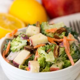 A white bowl filled with broccoli salad mixed with carrots, apple chunks, and nuts. A halved lemon and a red apple are in the background.