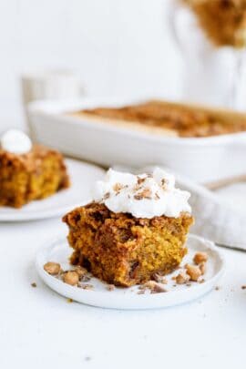 A slice of pumpkin cake topped with whipped cream and nuts is served on a white plate, with a baking dish and another slice in the background.