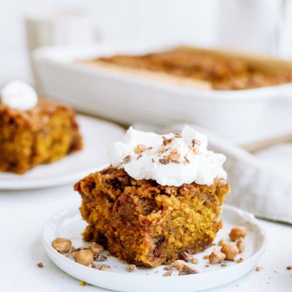 A slice of pumpkin cake topped with whipped cream and nuts is served on a white plate, with a baking dish and another slice in the background.