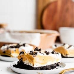 A slice of layered dessert with a crumbly dark crust, creamy filling, and whipped topping on a white plate with a gold fork. More slices and wooden cutting boards are blurred in the background.