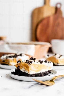 A slice of layered dessert with a crumbly dark crust, creamy filling, and whipped topping on a white plate with a gold fork. More slices and wooden cutting boards are blurred in the background.