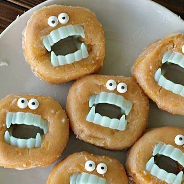 A plate of glazed donuts is decorated with plastic eyes and fake blue vampire teeth, creating a playful monster-themed design.