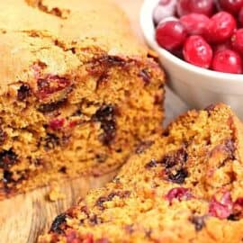 A loaf of cranberry bread with two slices cut and a bowl of fresh cranberries beside it. The bread is speckled with cranberries and appears moist and freshly baked.