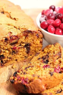 A loaf of cranberry bread with two slices cut and a bowl of fresh cranberries beside it. The bread is speckled with cranberries and appears moist and freshly baked.