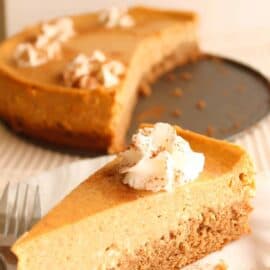 A slice of pumpkin cheesecake topped with whipped cream and a sprinkle of cinnamon is placed on a white plate with a fork. A partially cut cheesecake is in the background.