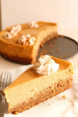 A slice of pumpkin cheesecake topped with whipped cream and a sprinkle of cinnamon is placed on a white plate with a fork. A partially cut cheesecake is in the background.